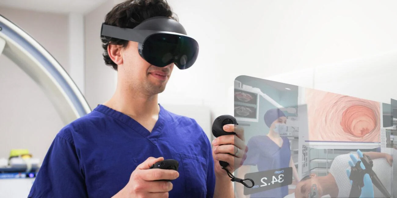 Person in blue scrubs wearing a VR headset and holding controllers beside an endoscopy simulation display.