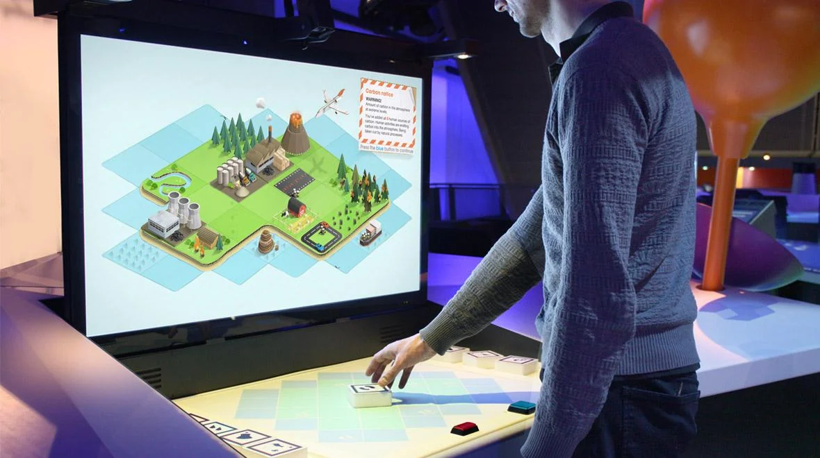 Visitor using a tabletop control beneath a screen showing the Carbon Cycle AR landscape.