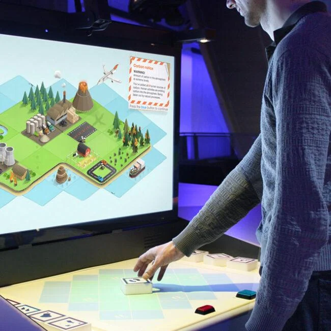 Visitor using a tabletop control beneath a screen showing the Carbon Cycle AR landscape.