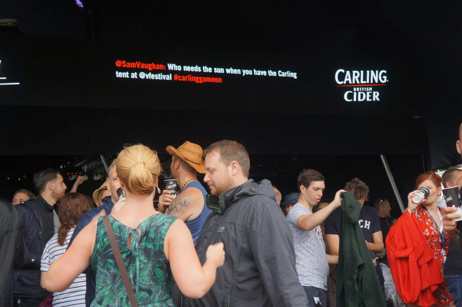 Festival bar visitors beneath a screen displaying a social media message and Carling Cider logo.