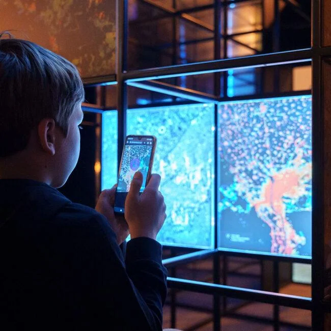 Visitor viewing a blue-lit Direction Earth/Space display through a smartphone.