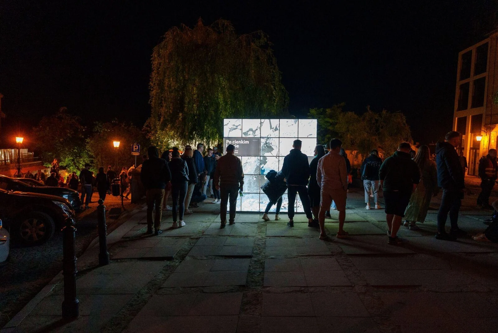 Visitors standing around illuminated exhibition panels in an outdoor square at night.
