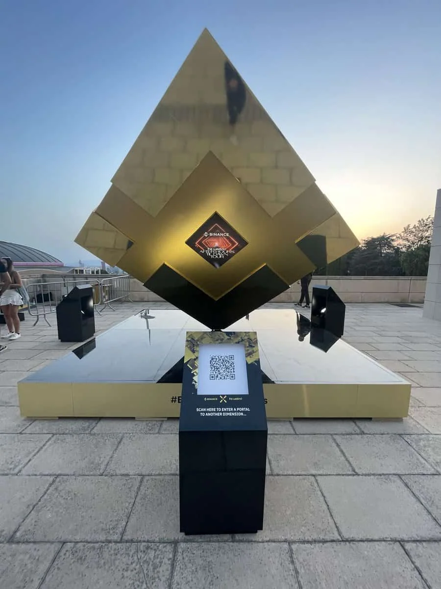 Gold geometric Binance sculpture with a QR-code pedestal in an outdoor plaza.