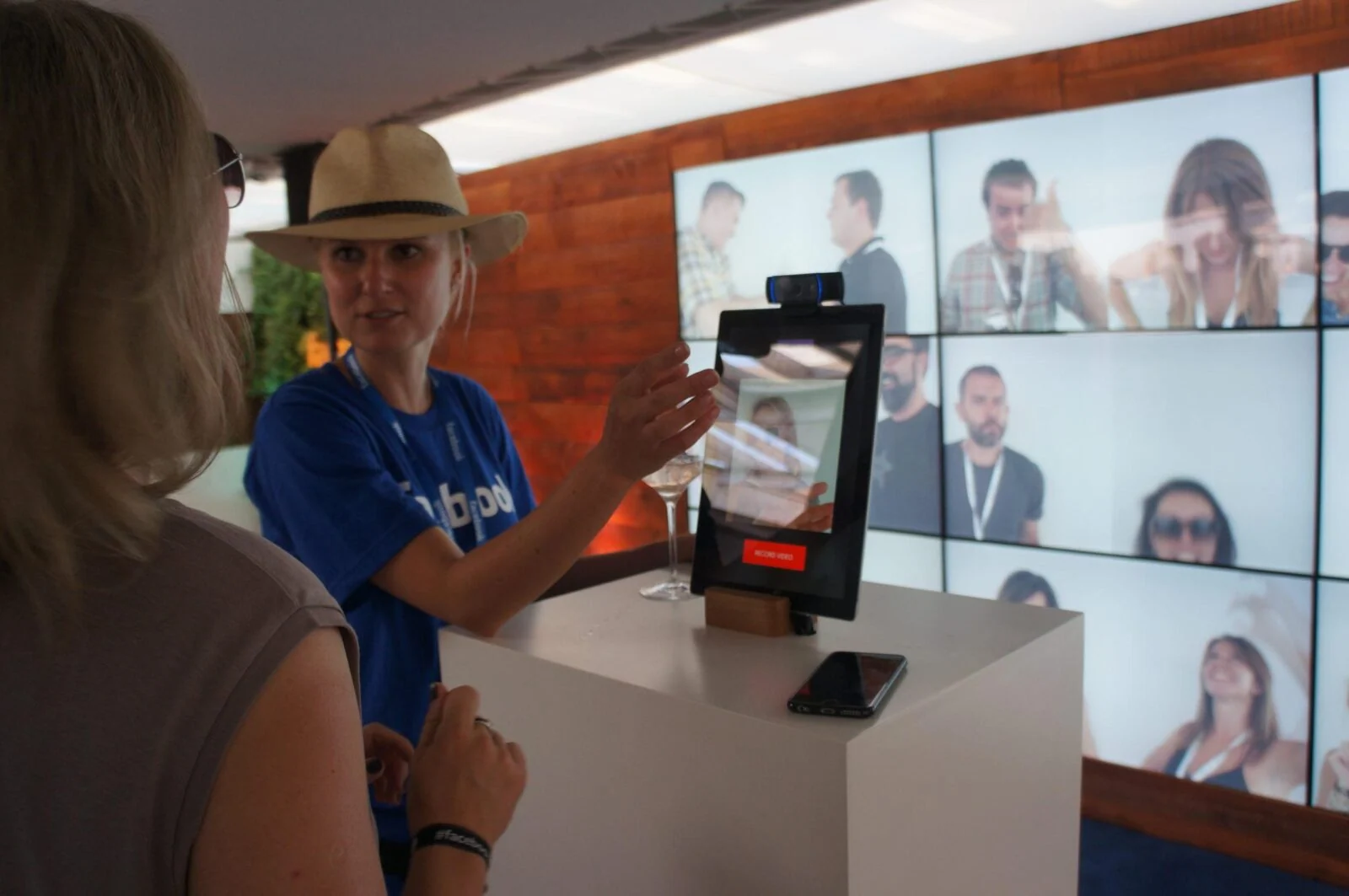Visitor and staff member using a camera kiosk in front of a wall of video portraits at Facebook zone.