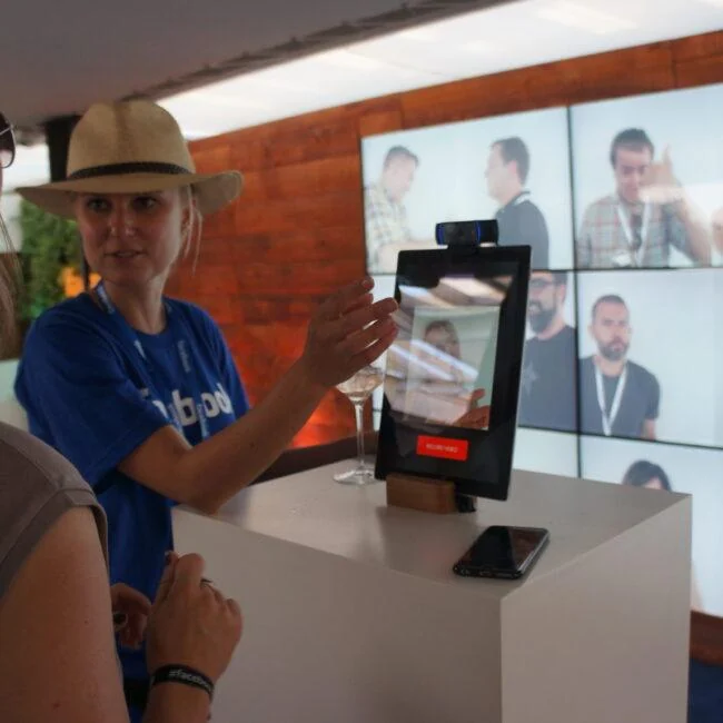 Visitor and staff member using a camera kiosk in front of a wall of video portraits at Facebook zone.