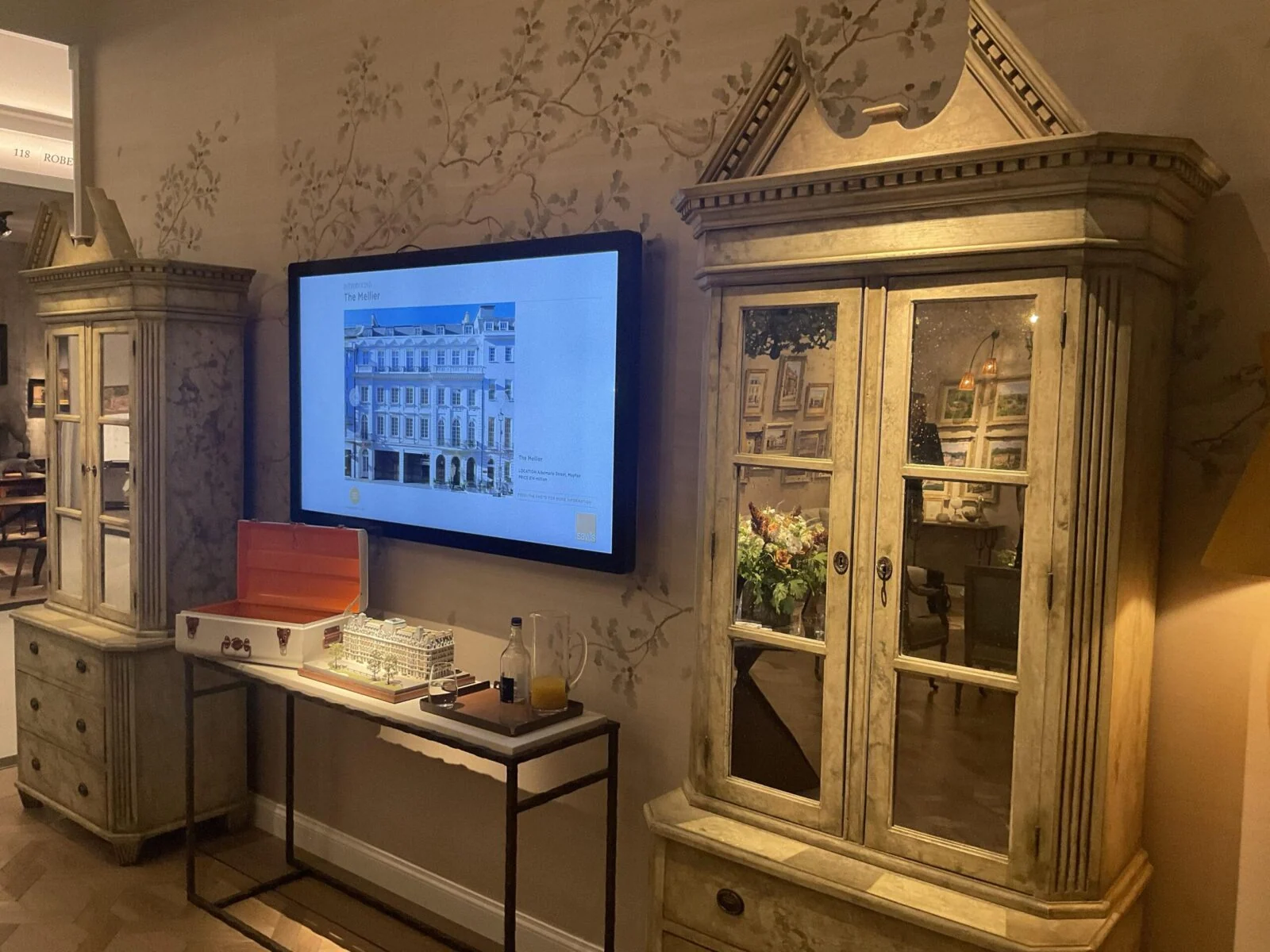 Architectural image on a wall-mounted screen above miniature buildings at Masterpiece fair.