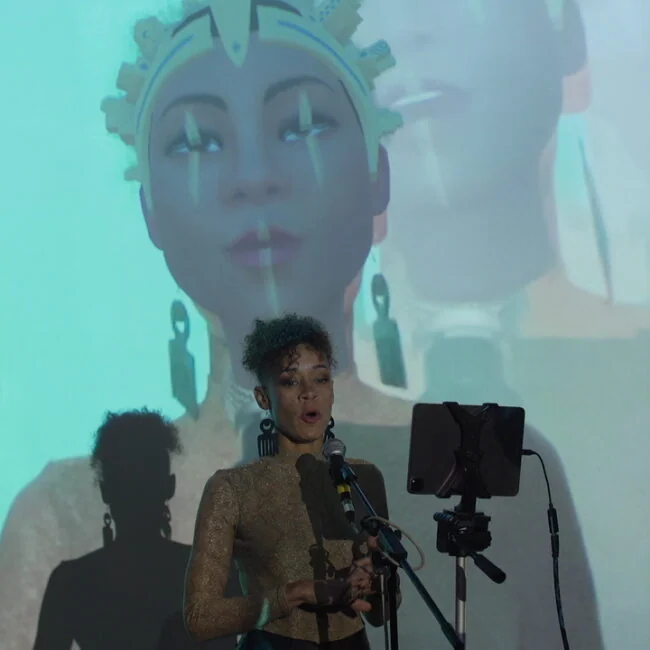 Live performer at a microphone with a large projected digital avatar behind them at Metamirror.