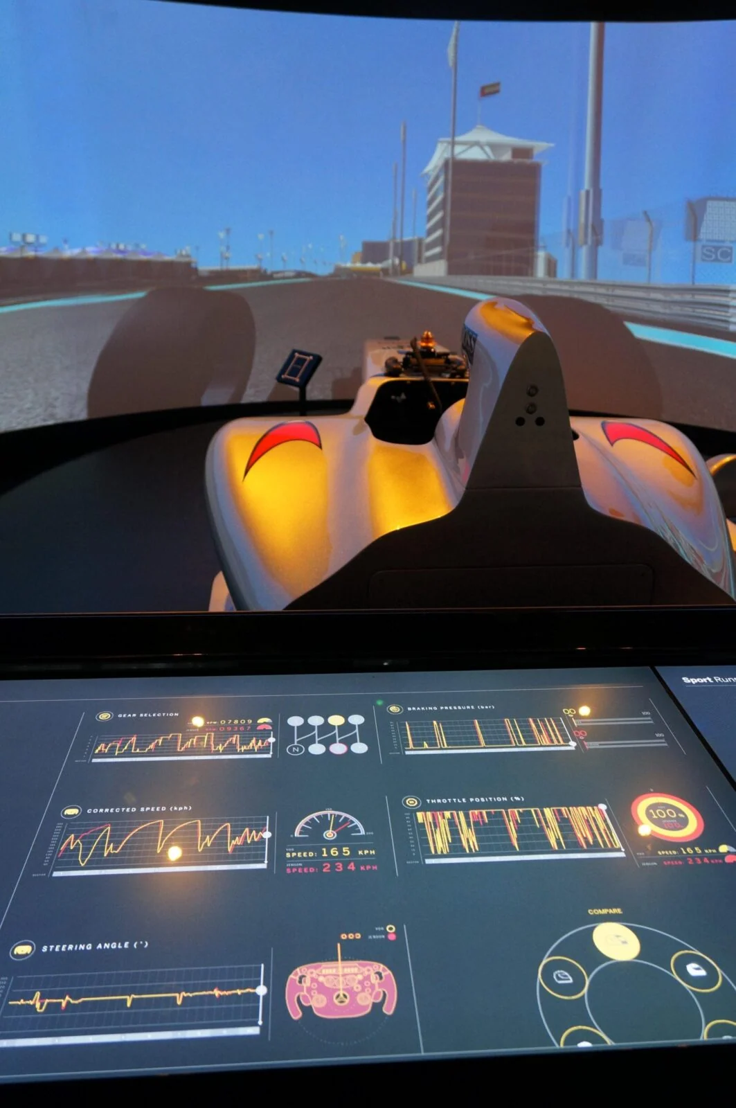 Panoramic racing display showing an orange car, with a touchscreen control panel below.