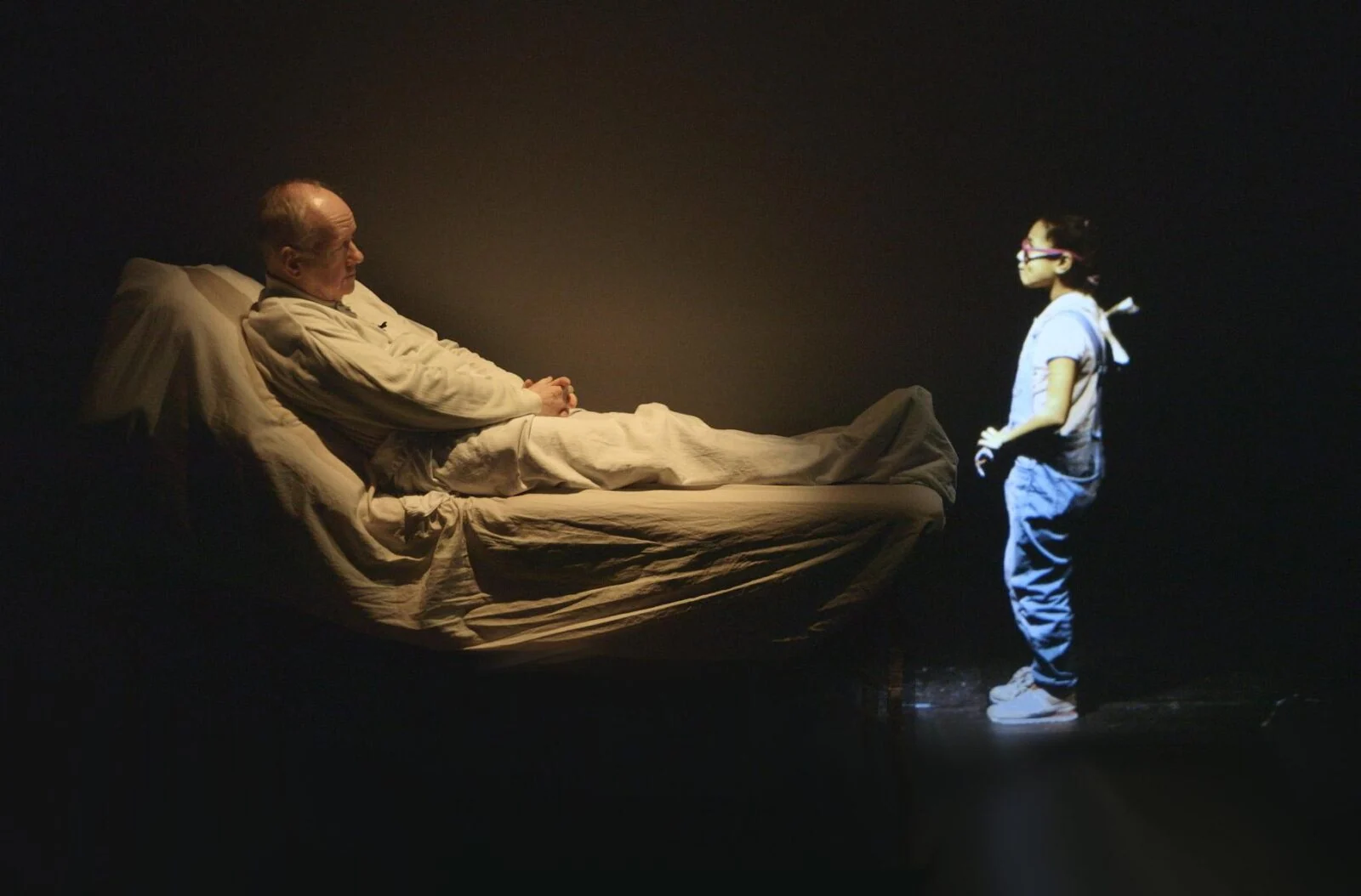 Performer reclining on a bed opposite a projected standing figure on a dark stage.