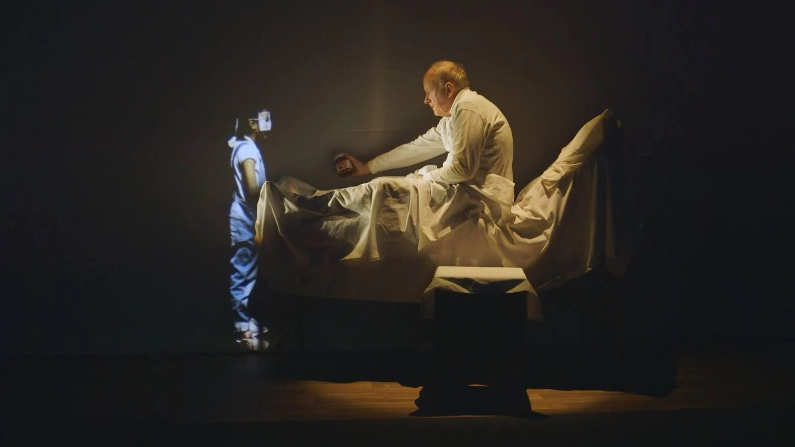 Performer reaching from a bed towards a projected character on stage.